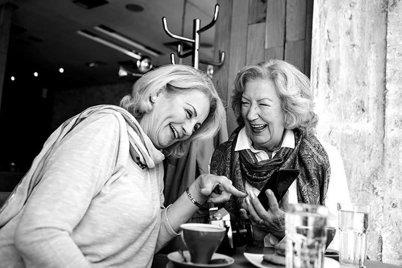 Two senior women sitting in modern cafe and drinking coffee, chatting in relaxed atmosphere, using smart phone