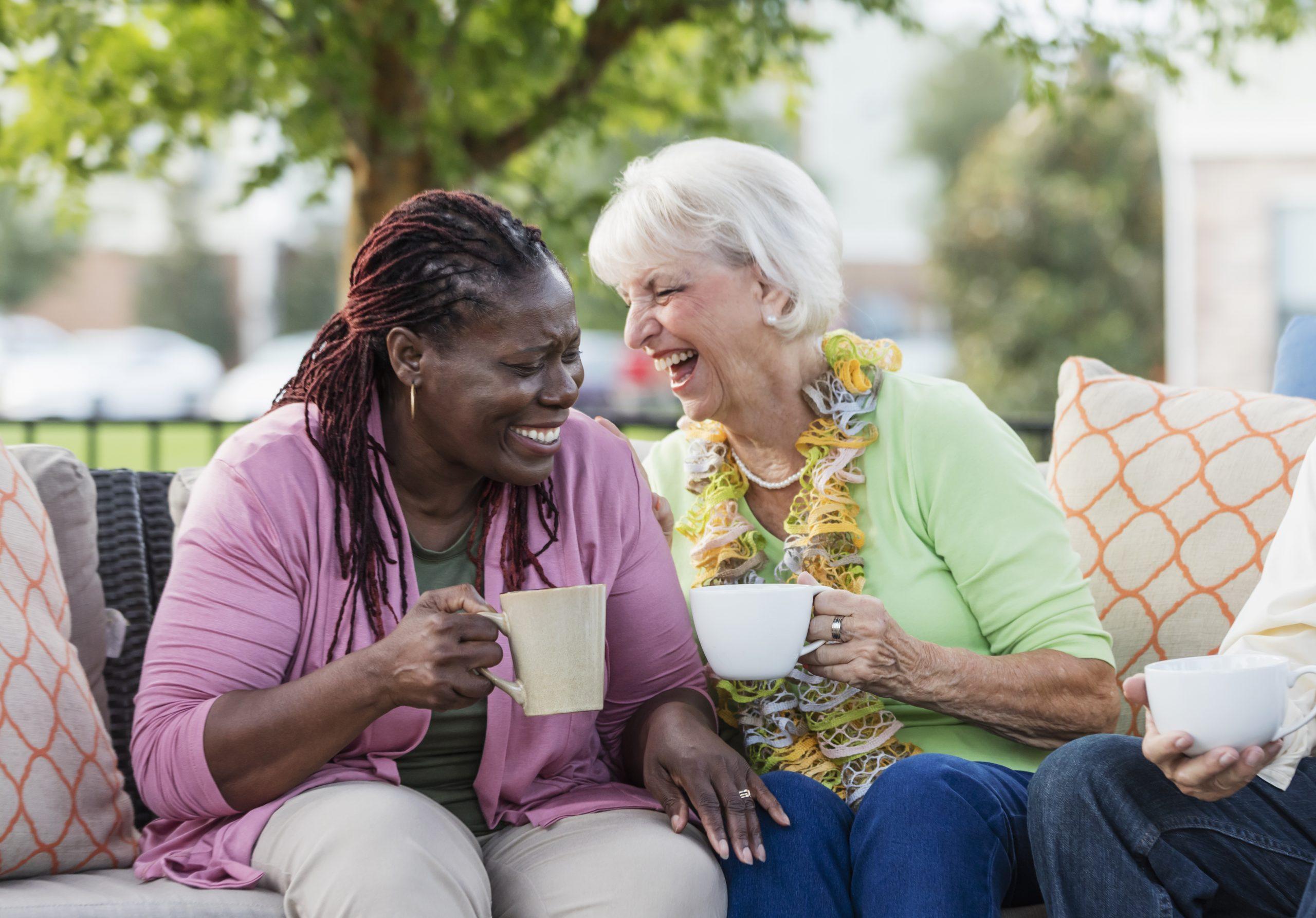 A senior woman in her 70s, drinking coffee and laughing with her African-American friend, a mature woman in her 50s. They are sitting outdoors on a patio sofa.