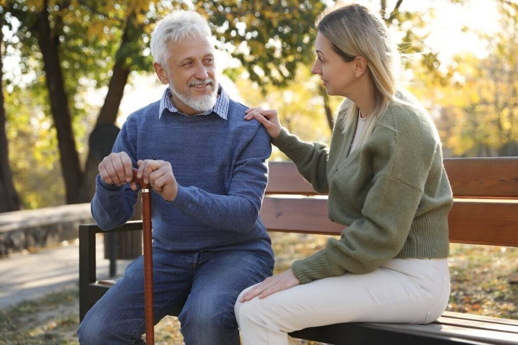 Caregiver assisting senior man on wooden bench in park.