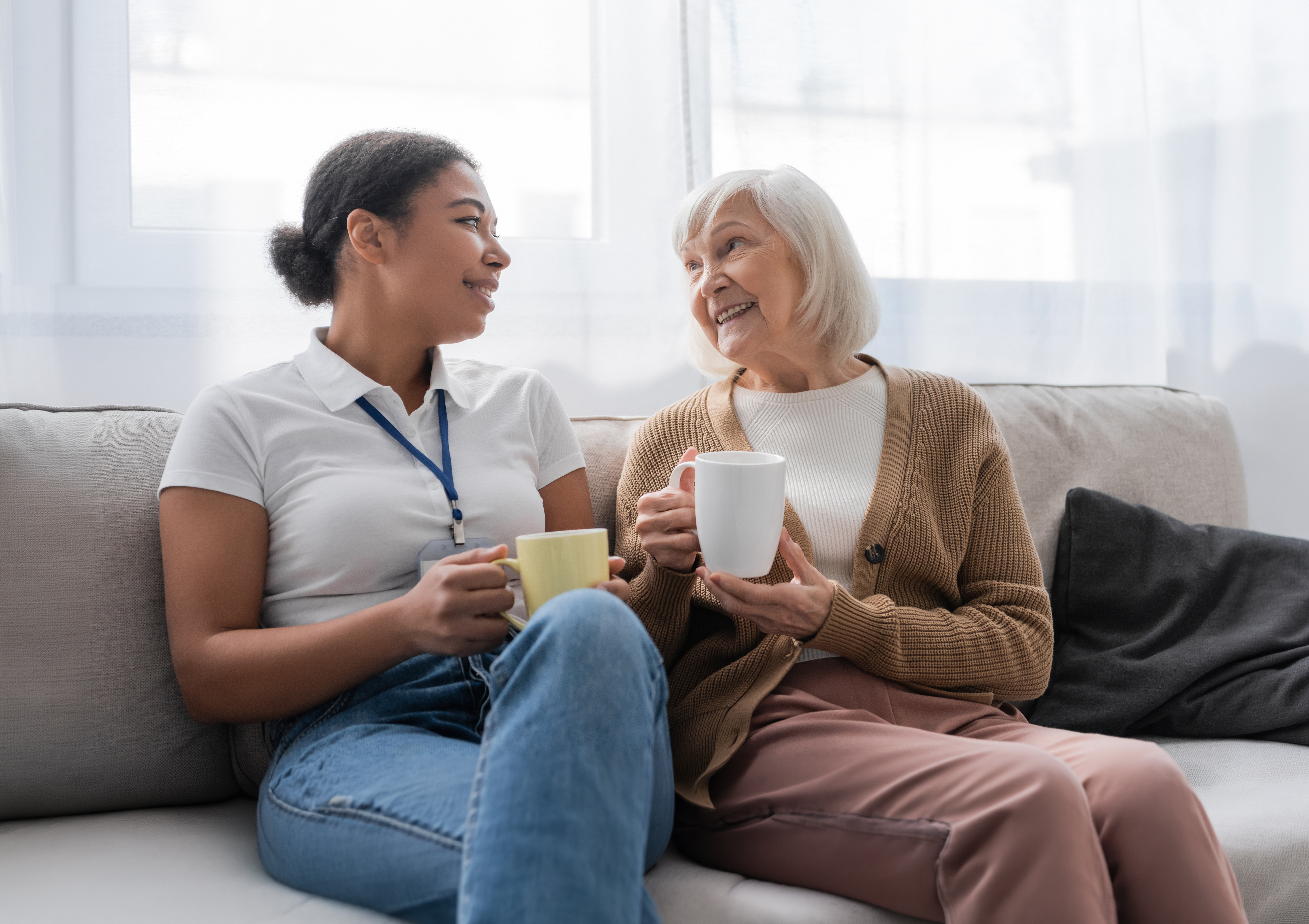 Two women sitting on a couch sharing a conversation and holding mugs in a bright room.