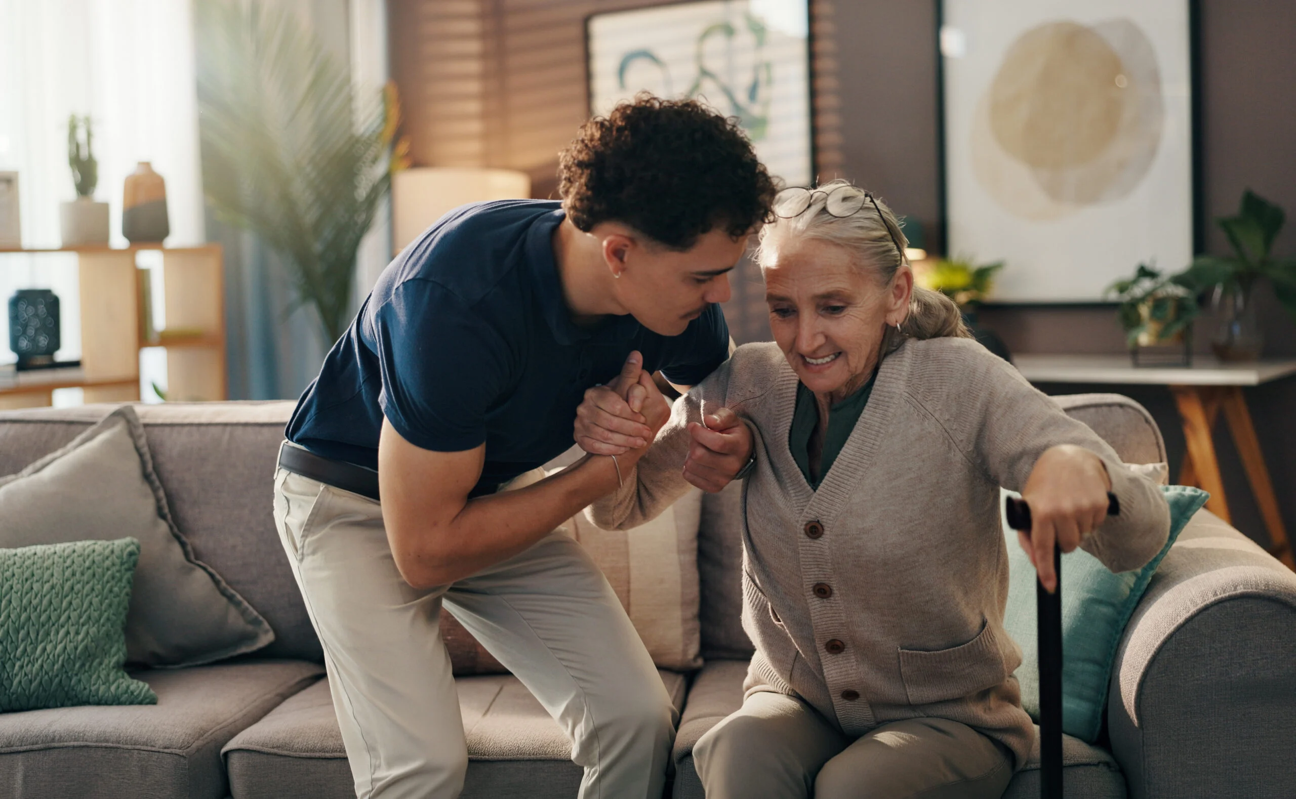 Younger person helping an older person with a cane stand up from a couch in a living room.