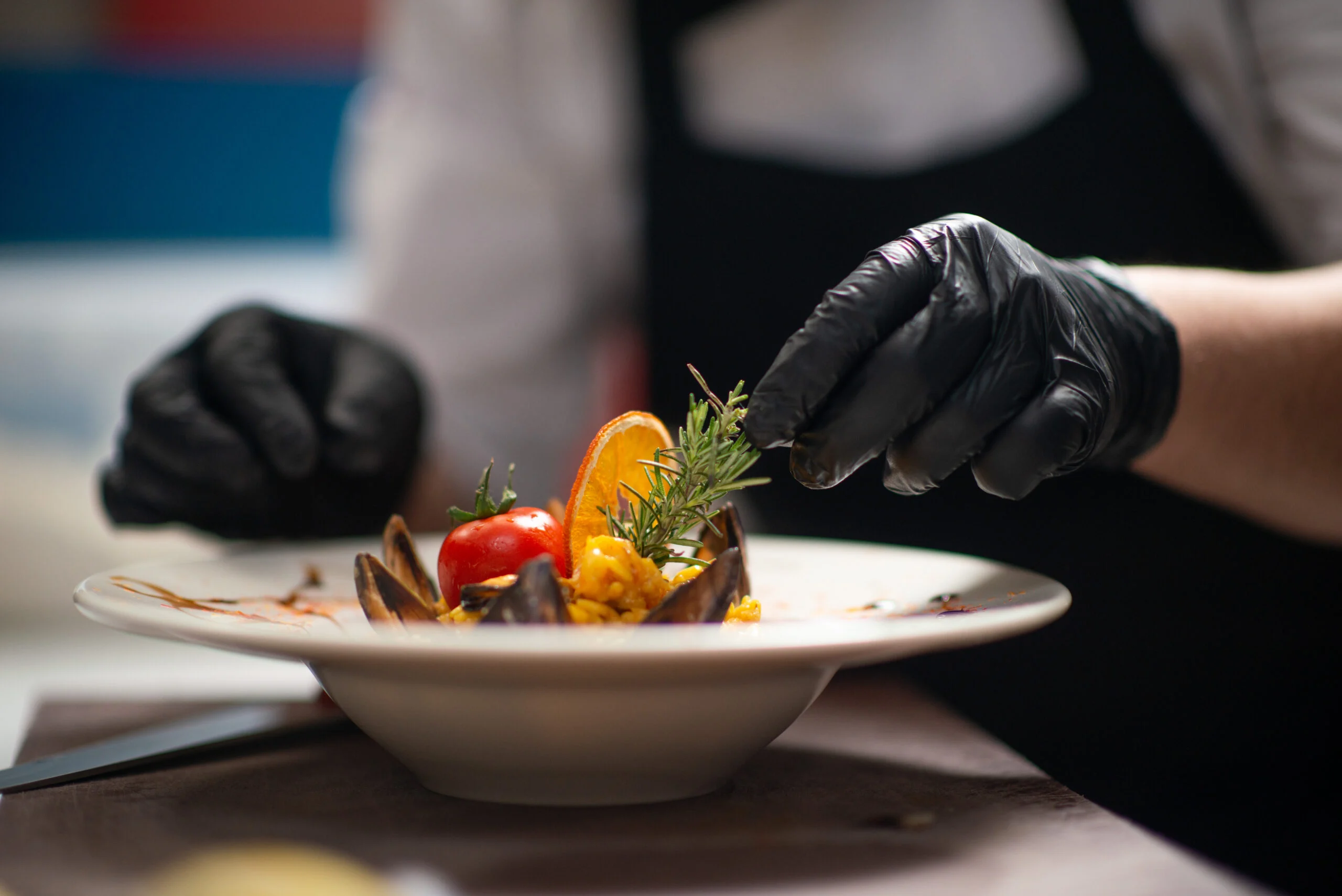 Hand wearing black gloves garnishing a plated gourmet dish with fresh herbs in a dining setting.